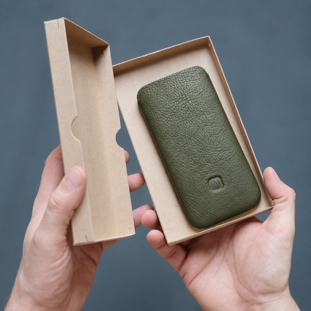 in einem Produktkarton, das von Männerhänden gehalten wird, liegt eine olivgrüne Handyhülle aus Leder